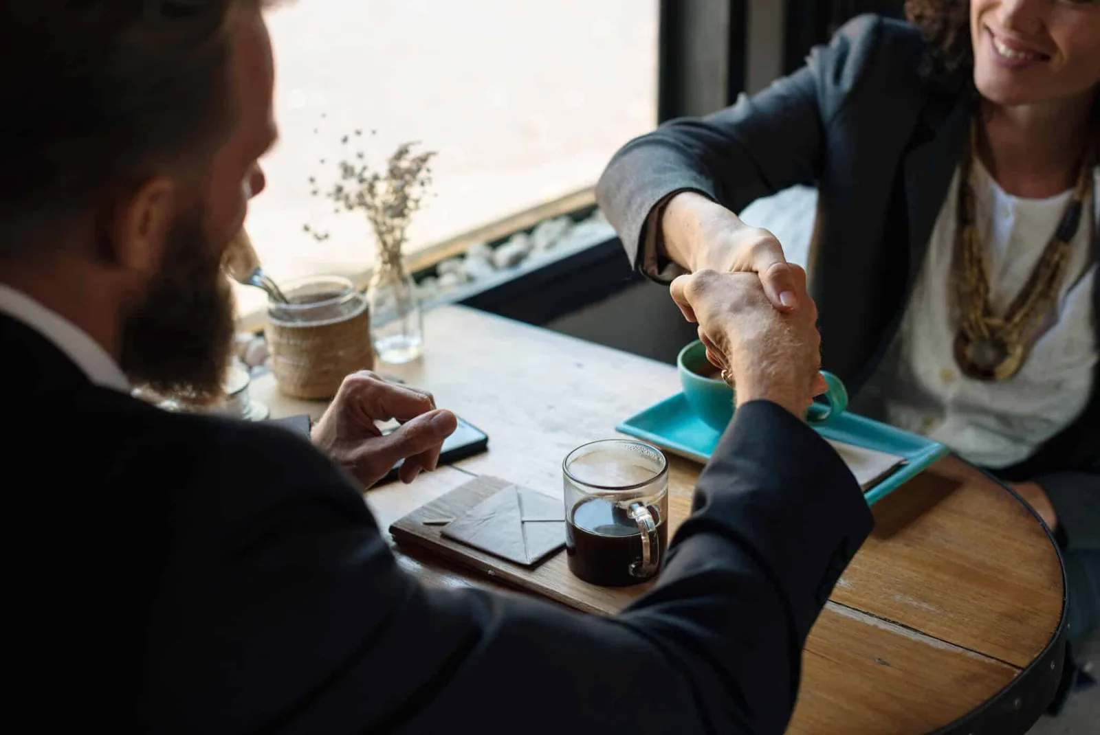 seo_kontaktaufnahme Zwei Geschäftsleute geben sich am Cafétisch mit Kaffee die Hand, Symbol für SEO-Kontaktaufnahme